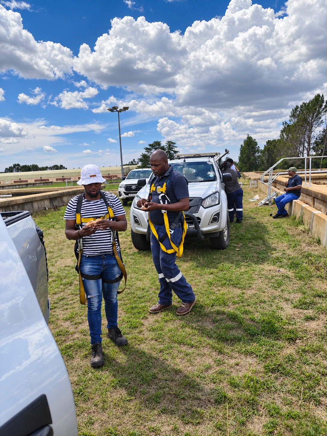 Two team members in safety harnesses on a grassy site beside a bakkie